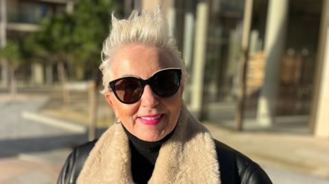 Kim Carpenter is a woman with short white hair. She is smiling into the camera. She is wearing a black turtle-neck and a black aviator coat with a fluffy collar. She has big black sunglasses on and pink lipstick. The background is an out-of-focus building at the waterfront with a paved path around it. 