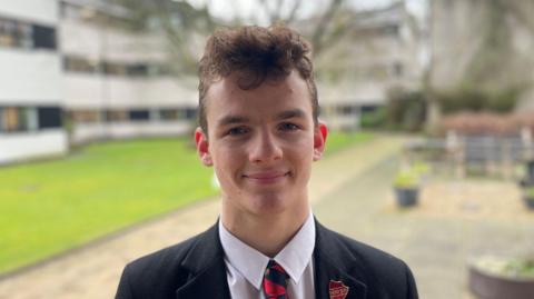 Oscar Scullion has short brown curly hair. He wears a black blazer and white shirt with a red ad navy striped tie. He has a red badge in the shape of a shield on his lapel. Out-of-focus, behind him is a green garden with a white, multi-storey building behind it.