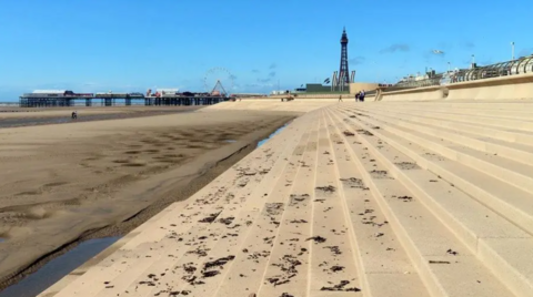 Steps leading on to Blackpool beach, with the tide out. The tower and pier can be seen in the distance against a blue sky