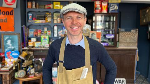 Anthony Bates stands in his barber shop in Bridlington. He is wearing a beige apron, a blue jumper and a flat cap. In the background is a display of 1940s memorabilia including telephones, furniture, groceries and ornaments.