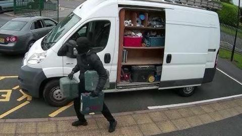 A man in a black coat carrying three toolboxes away from a white van with its doors open.
