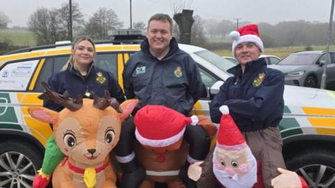 One woman and two men wearing dark jackets and in inflatables costumes, looking like they are the backs of a reindeer, gingerbread man and a Santa. The people all standing in front of a yellow and white car