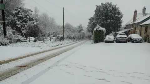 A road is covered in snow with tyre tracks. Cars are under a blanket of snow