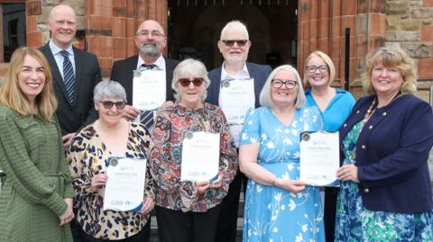 Three men and six women stand should to shoulder in two rows outside the red brick building of Derry's Guildhall. They are being presented with certificated.