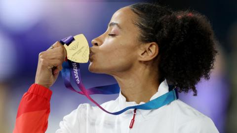 Jazmin Sawyers is sideways on to the camera as she kisses her medal, in her right hand. She has tied-back black frizzy hair and is wearing a white team top.