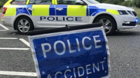 Police sign reading "police accident". Behind the sign is a white police vehicle with yellow and blue check patterning.