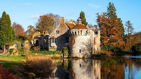 A pretty English country house. It is surrounded by trees and there is a pool of water in front of it. 