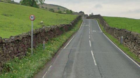 A country road runs between two fields, one of which has a sheep in. There is a 30mph sign at the side of the road.