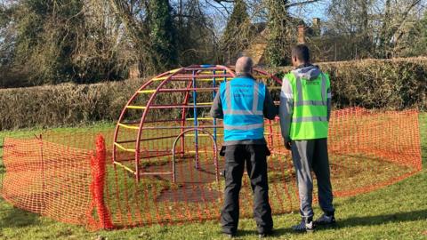 Two men one in a blue hi-vis jacket and one in a yellow hi-vis jacket looking at a fenced off climbing frame