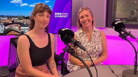 Lucy Sheppard and Aimee Peach sit next to each other in the BBC Radio Somerset studio. Lucy is wearing a black top and a skirt with a flowery pattern and Aimee is wearing a black and white dress. The studio is lit up in purple colours.