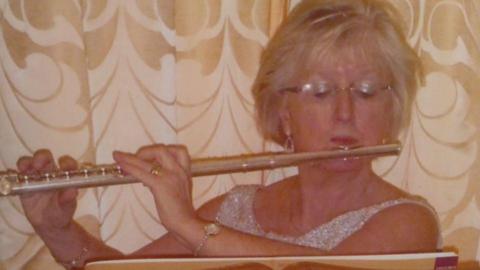 An older woman with mid-length blonde hair and glasses looks down as she plays the flute and reads sheet music. She is wearing a sleeveless glittery silver dress and is wearing a dainty watch on one wrist and a bracelet on the other. She is stood against a ruffled gold and white curtain.