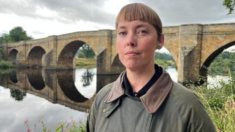 Sophia Stovell standing in front of the Chollerford Bridge, a long stone structure with five arches, over the River Tyne. She is standing on the river bank at the George Hotel in Chollerford. She is wearing a brown and green jacket with a black shirt. Her hair is light brown and has a short fringe. 
