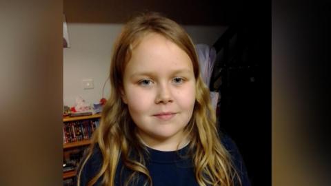 A girl wearing a black top looks at the camera and smiles with no teeth.