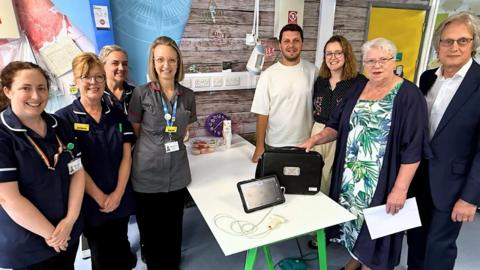 Staff at Royal Surrey’s Onslow Ward pictured to the left of the donated ultrasound machine, with family and friends of Matt Long pictured to the right. The ultrasound machine looks like an iPad and is accompanied by a briefcase.