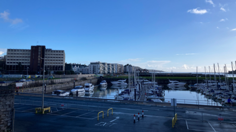 A marina. Boats are docked in the marina. The sky is blue. Tall buildings are on the left hand side.