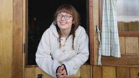 A woman wearing a white jacket and glasses. Her hair is in two plaits. She's smiling and looking outside a window in a wooden building.
