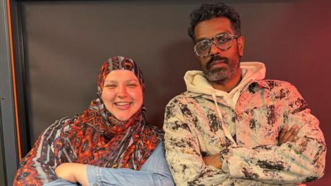 A woman wearing an orange, patterned headscarf smiles at the camera. She stands next to a man with short, curly hair wearing glasses. He looks at the camera. They both have their arms folded.