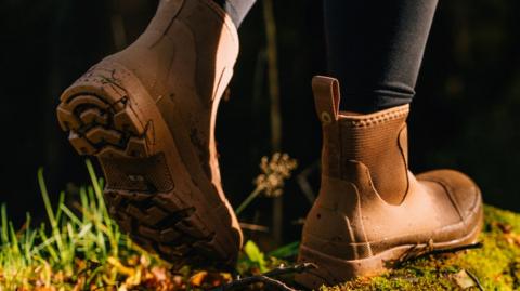 A pair of feet in brown ankle-length boots walk along what appears to be moss-covered log. Blades of grass can also be seen. The person's legs can be seen in black leggings.