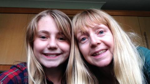 A young girl smiles in a selfie with her mother, in front of kitchen cupboards. Both have blonde hair.