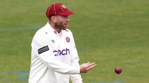 Luke Procter throws a red cricket ball to an unpictured player