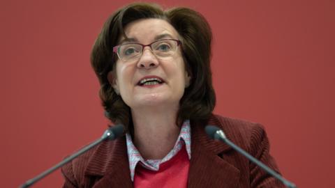 Eluned Morgan is stood looking up from a lecturn which has microphones on top of it. She is wearing a dark red jacket and a red top with a shirt underneath.