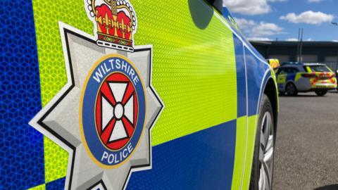 The side of a marked Wiltshire Police car, which is parked in a car park. 
