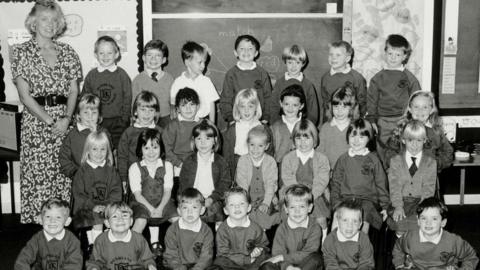 A black and white photo of Gwen Mayor, a blonde-haired woman in a patterned dress, and her 28 pupils arranged in four rows of seven in a classroom. The children are in school uniform.