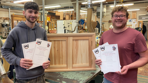 A picture of Alex Gill on the left with Gareth Foster to the right. They are both holding their certificates and are stood in front of a wooden stand.
