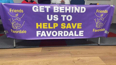 A purple sign with the words "Get behind us to help save Favordale" in white and yellow lettering. The banner has the Friends of Favordale logo on either side.