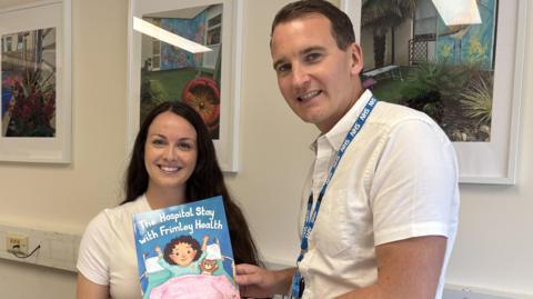 A woman with long dark hair is standing next to a man with dark hair, both are wearing white tops. The man is holding a copy of a book called The Hospital stay with Frimley Health