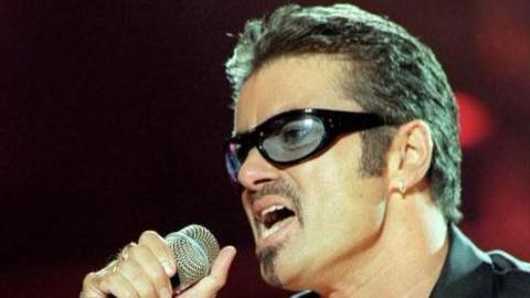 George Michael performing for NetAid at Wembley Stadium. He wears tinted glasses and has a short goatee beard.