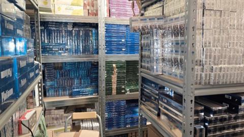 Metal shelves in a room and absolutely full of packets of cigarettes. 