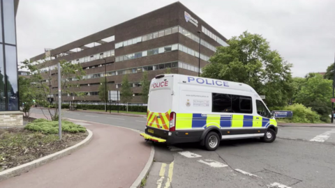 A large police van is parted across the road on the edge of a roundabout. Behind it is a large brown brick building with windows along each floor. Trees fringe the road ahead. To the left is the corner of an entirely glass building, the glass appearing black and reflecting the surroundings. On the ground in front of it is some scrubby soil.