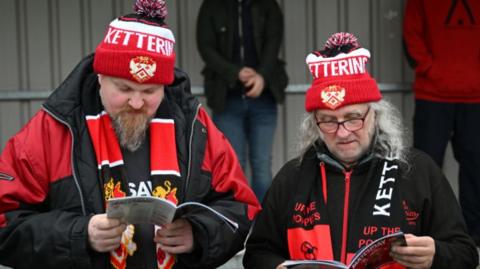 Fans of Kettering Town read the match day programme prior to the Emirates FA Cup Second Round match between Kettering Town and Doncaster Rovers at Latimer Park