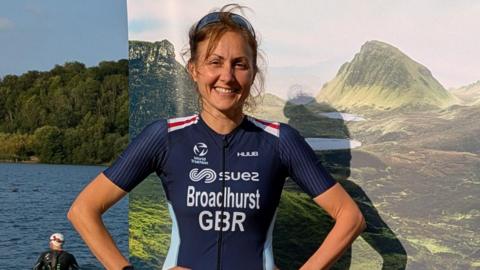 A woman wearing a dark blue Team GB outfit, has her hands on her hips and smiles while standing in front of a poster, with a lake behind it.