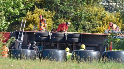 the underside of a lorry can be seen after it overturned. there are a number of rescue workers on top of the lorry and standing to its side. The area is surrounded in green bushes, trees and long grass. 