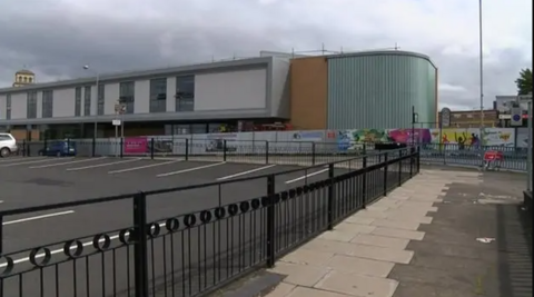 The youth club building is curved at one end with a light blue-green facade. The other part of the building to the left is grey with large windows. There is a car park in front of the building, with railings in the foreground of the shot.