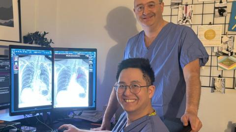 Consultant radiologists Dr Daniel Fascia and Dr Andrew Koo, who wear blue scrubs, sit and stand next to a computer showing a chest x-ray.