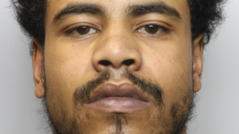 A close-up mugshot of a young man with black frizzy hair, a sparse beard and dark eyes. He has a neutral expression. 