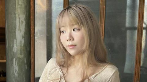 A woman with mid-length blonde hair and a fringe looks to her right, wearing a beige top and winged eyeliner.