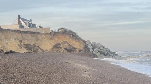 The Thorpeness coastline that has been eroded away. Large cliffs can be seen where the erosion has happened. Rock sea defences have been put around the cliff in the distance. A house above the cliff can be seen.