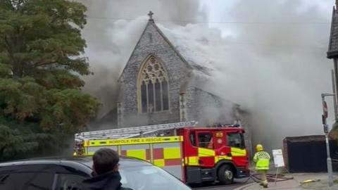 A Victorian flint-built church covered in smoke. In front of it is a red and yellow fire engine with a yellow-dressed fire fighter walking towards it.