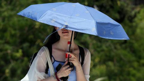 A young woman shades herself by holding a blue umbrella. She is also holding a bottle of cola.