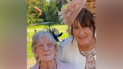 Beverley wears a smart white blazer, a beaded necklace and a pink fascinator hat. She has brown hair. On the left is her mother, wearing glasses, a pink dress or blazer, and appearing with white hair. It is sunny outside.