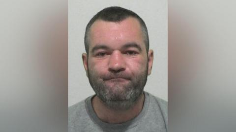 Mugshot of a man in a grey sweatshirt with short dark hair and a grey stubbly beard