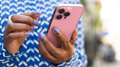 Close-up of a pair of hands with purple painted nails holding a pink phone. The person is wearing a blue and white print shirt. 