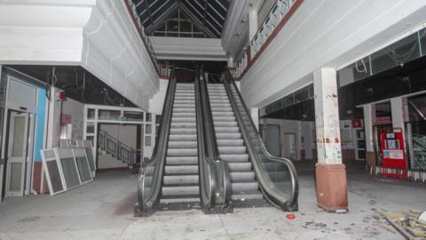 A pair of immobile escalators in a deserted shopping centre, with rubbish littering the floor.