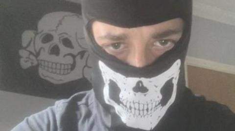 Alfie Coleman wearing a black balaclava with a skull on it. Only his eyes can be seen. Behind him a black flag with a white skull and crossbones is pinned to a wall.