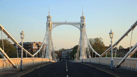 A view from the middle of the road across Albert Bridge as the sun comes up, illuminating the white and pink painted structure. There are buildings and trees in the background