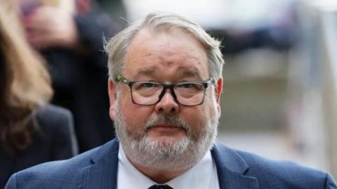 Close up of Christopher Mills outside court. He wears a blue suit, a white shirt and has black glasses. He has short grey hair and a grey beard.
